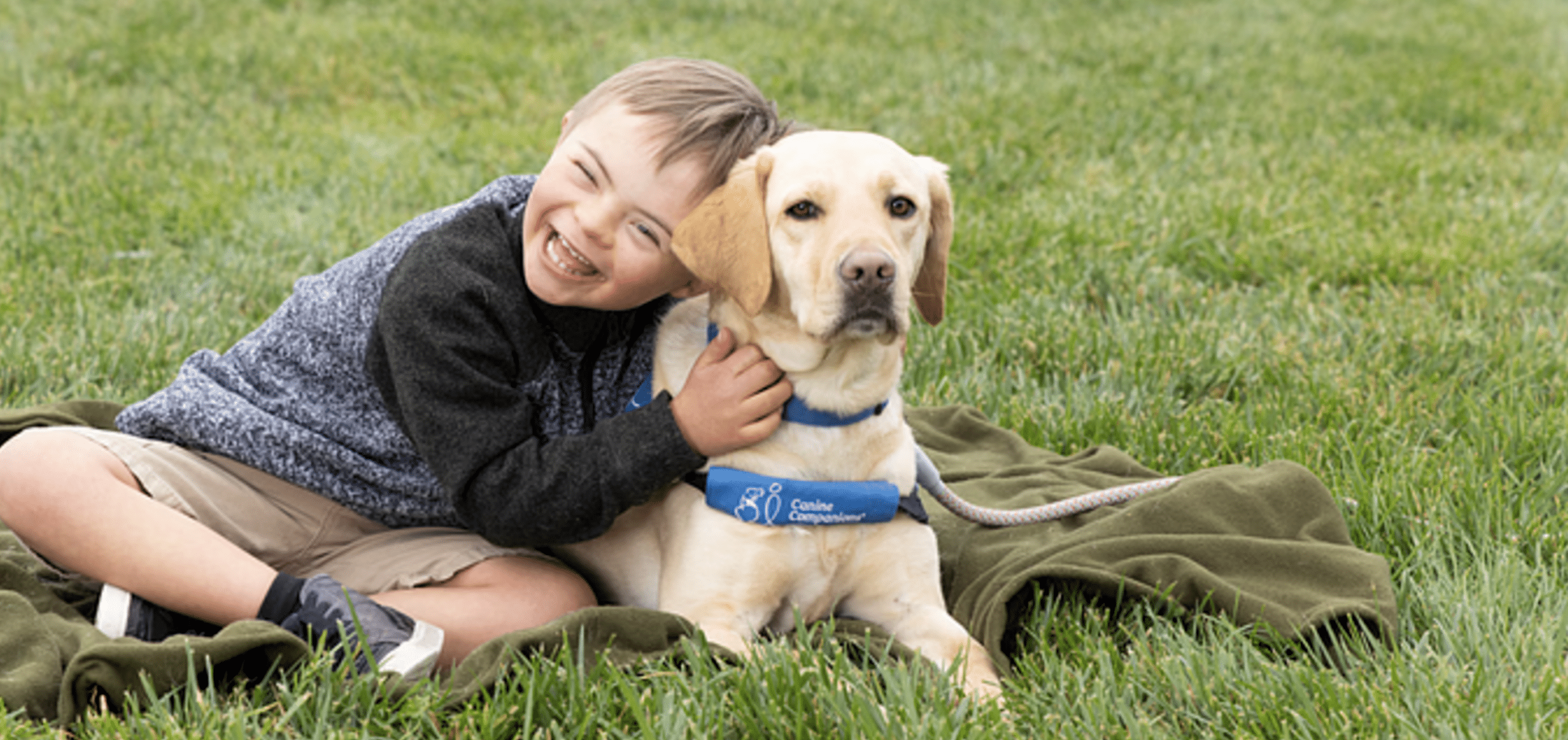 A child sitting on grass joyfully hugging a Labrador wearing a blue "Canine Companions" vest.